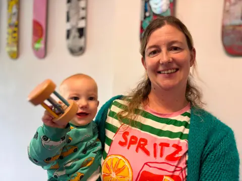 Smacking children could lead to lower GCSE grades, study suggests Joshua us on the left in a green jumper with cars on it and is being held by mum, Sarah, in a greed cardigan, green and white striped top with a drawn glass with red liquid and a slice of orange with the word 'Spritz'