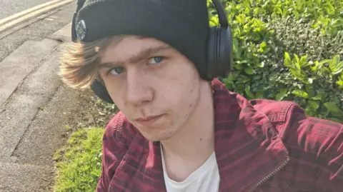 Joshua Clover is wearing headphones with a black beanie. He has on a red jacket.