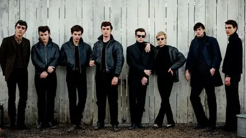 Gordon Timpen/ Hamburg Films Ltd The band's cast poses with actors Luna Jordan and Casper von Bülow against a large fence. They are all wearing black outfits.
