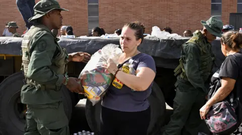Reuters Venezuelan military personnel distribute aid to people affected by deadly earthquakes that hit the country. In the middle of the image, a woman carries a package of food over her shoulder.