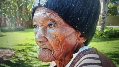 The Polygamist's creator says women see themselves reflected in her Netflix hit Sue Nyathi family An elderly woman wearing a black knit beanie and a brown patterned sweater sits outdoors in a sunlit garden. The image is a close-up side profile, highlighting deep facial lines and a thoughtful expression as she looks into the distance. Lush green grass, trees, and leafy plants fill the softly blurred background, creating a peaceful natural setting.