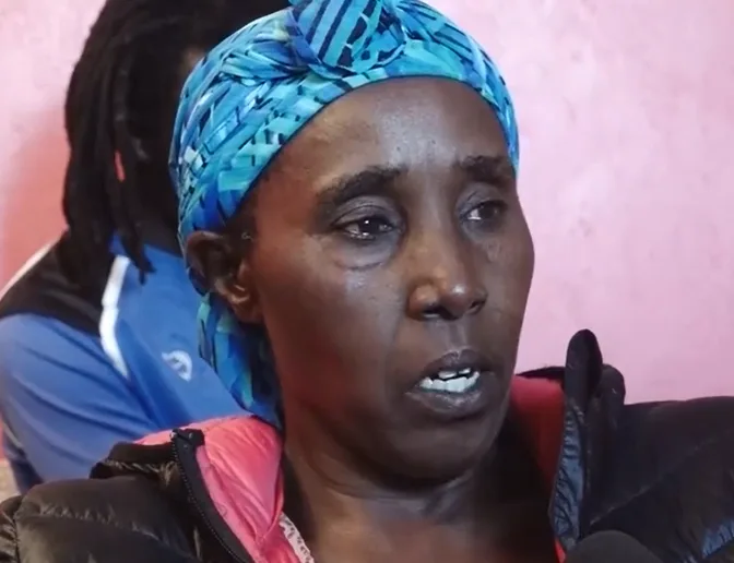 Sje is wearing a turquoise patterned headwrap and a black coat wth a red-lined hood. her eyes are a bit teary. behind her, is a man, whose face is blocked by her head.