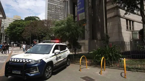 Getty Images A police car with its lights on is seen parked on a pavement outside the library - a large grey building - as pedestrians walk in the background. A police officer with his arms crossed is seen stood next to the car