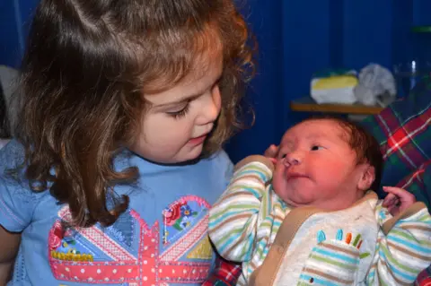 Kate looks down at newborn James being held in patterned clothing.