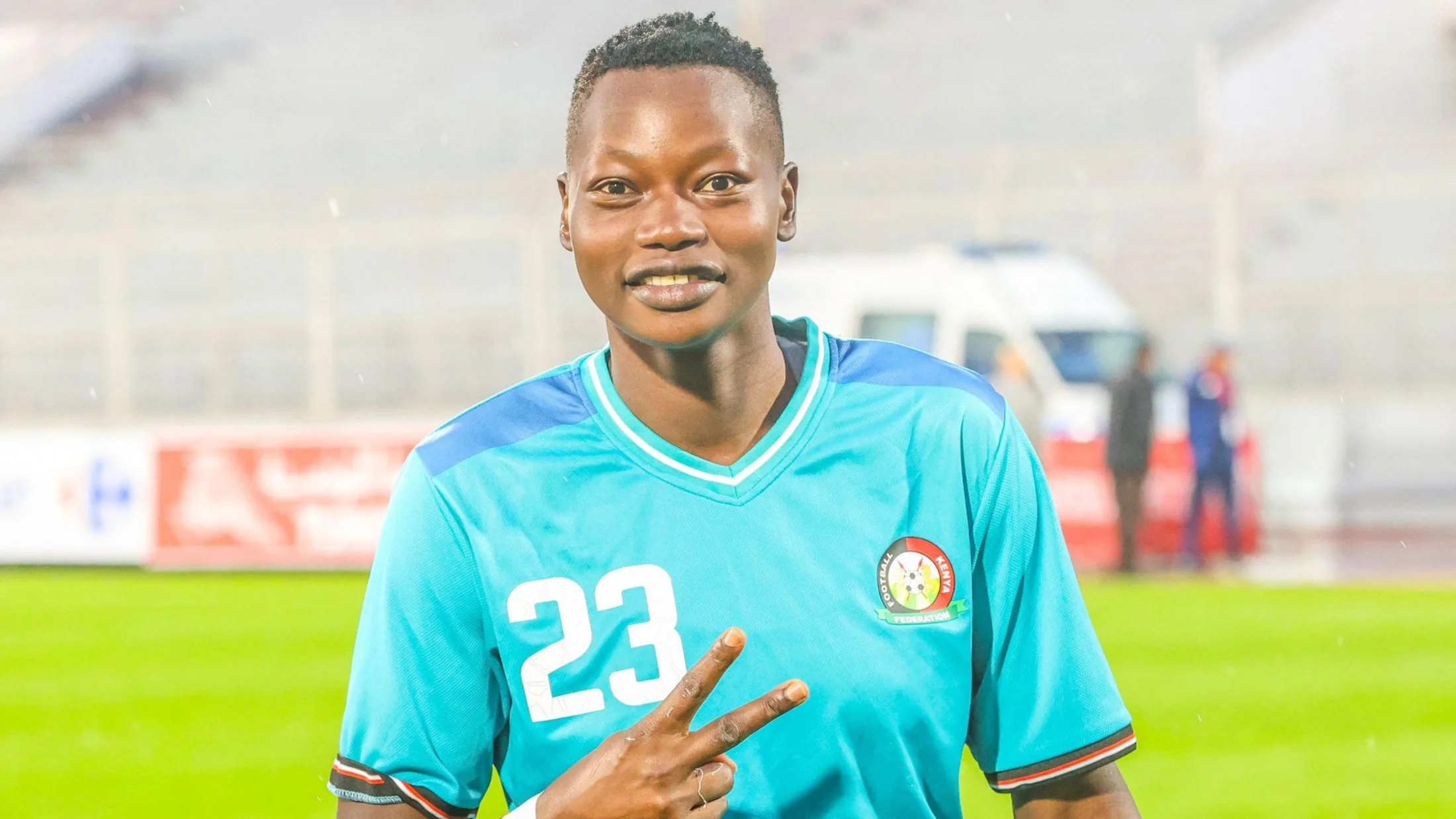 Lilian Awuor, visible from stomach up wearing a turquoise Kenya goalkeeping shirt with a white number 23 on the right breast, holds up two fingers at a roughly horizontal angle to the camera while smiling. An empty green pitch and stand are blurred in the background, as is an emergency vehicle positioned just behind advertising hoardings, with two men standing near the front of the vehicle