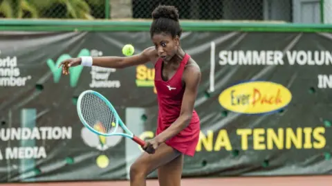 William Klu A young black woman playing tennis wearing an all red outfit, holds a green tennis racquet.