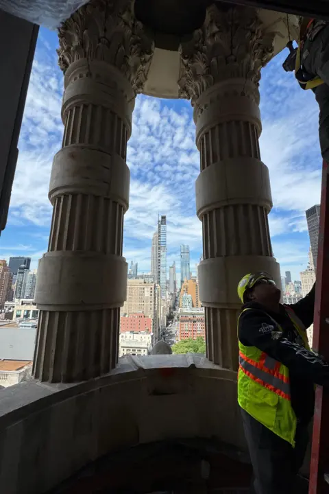 Would you pay $58.5m to live in this iconic New York building? Grace Eliza Goodwin Image of construction worker in yellow vest with a ladder on a balcony with large columns and a view of the Manhattan skyline