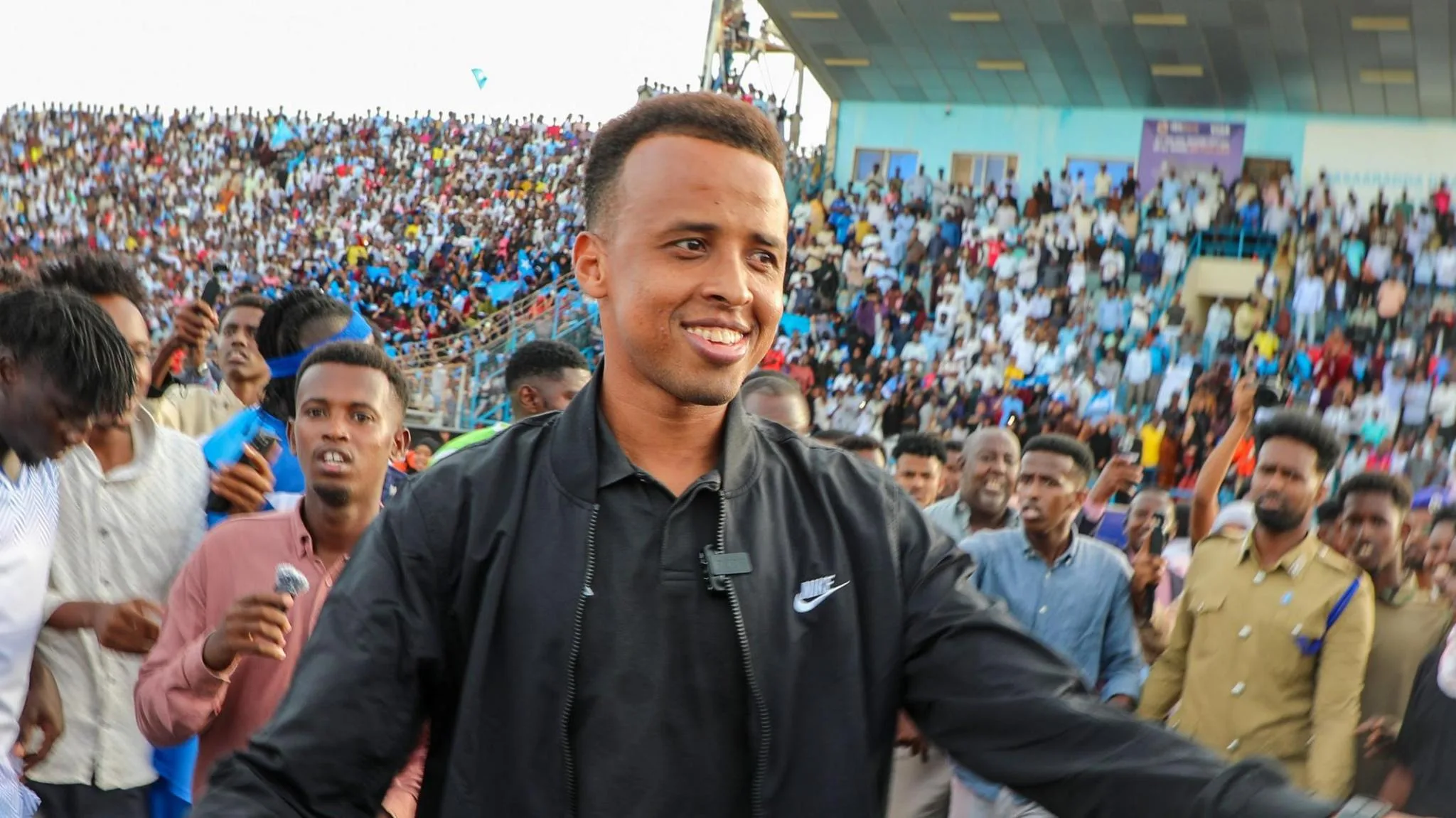Omar Artan, visible from stomach up and smiling while wearing a black tracksuit top over a black polo shirt, stands in front of a packed stand at a stadium as several people, including what looks like a member of security staff dressed in Khaki, mill around behind him