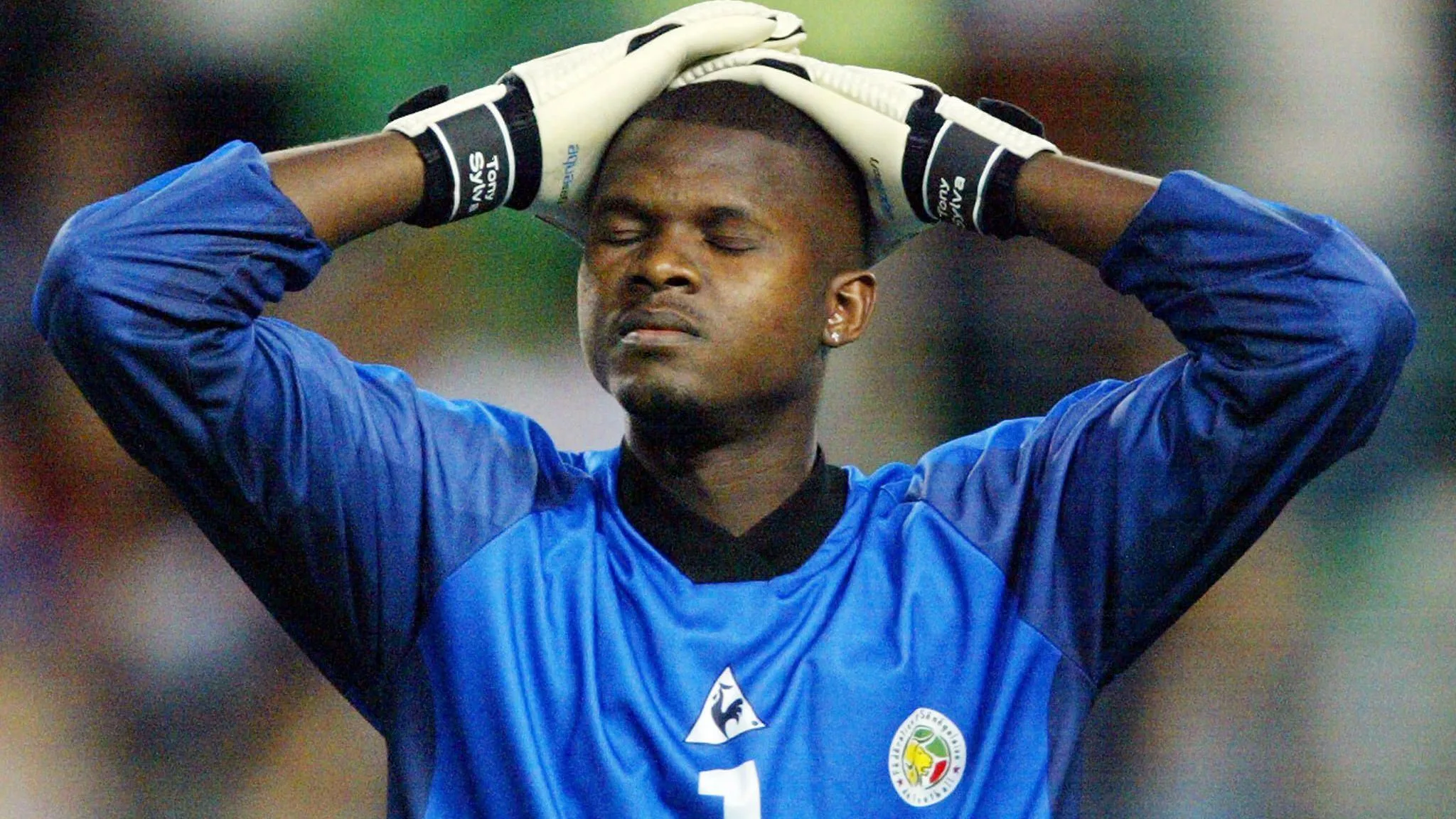 Senegal goalkeeper Tony Sylva, visible from chest up wearing a long-sleeved blue Senegal shirt, closes his eyes and holds his gloved hands to the top of his head with a look of frustration on his face