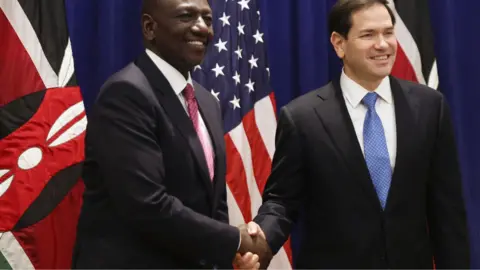 AFP via Getty Images Marco Rubio (R) and Kenya's President William Ruto (L) dressed in dark suits and ties shake hands in New York in September 2025 in front of the Kenyan and US flags.