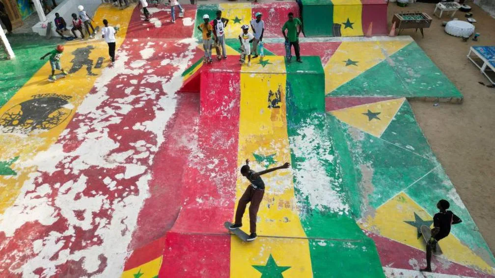 A high-angle wide shot of a bustling colourful skate park in the Ngor neighbourhood of Dakar, Senegal. The park is a vibrant hub of activity with skaters, either active on the ramps or stood watching. The entire concrete surface of the park, including curved transitions and steep ramps, is painted in the bold green, yellow and red colours of the Senegalese flag