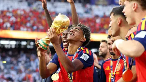 Michael Regan - FIFA / Getty Images Lamine Yamal lifts the golden World Cup trophy in the air as his teammates celebrate