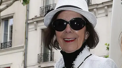 Getty Images Mary Jo Campbell - the mother of Kris Jenner smiles while wearing dark sunglasses, a white hat and blazer.