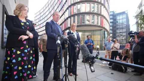 PA People stand behind microphones on a city street as a speaker addresses reporters outside a modern office building, while journalists and camera crews gather nearby.