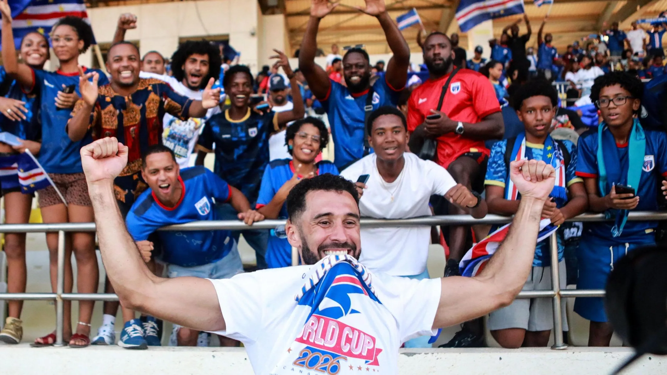 Roberto Lopes, visible from chest up in the foreground of the image, holds his arms up high as he celebrates Cape Verde qualifying for the 2026 World Cup. He wears a white T-shirt with the text World Cup 2026 on it, which he raises up by biting it between his teeth. Twelve joyful Cape Verde fans, mostly wearing blue team jerseys, are seen in the stands behind him, with other people and some Cape Verde flags seen out of focus in the distance