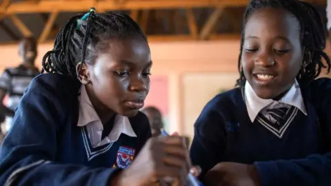 Dancing with Shakira at World Cup was 'very, very, very, very, very good' - Ghetto Kids AFP via Getty Images Two 13-year-old girls, who are members of the Ugandan dance group Ghetto Kids, attend school in Kampala on 20 Apri 2023. They are looking down at a book and wearing navy school jumpers and white shirts.