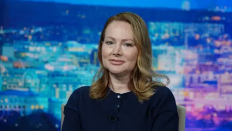 'He liked the fear in our eyes', Epstein survivors tell BBC Thierry Humeau Chauntae Davies, with blonde shoulder-length hair, wearing a navy jumper, looks straight ahead. A blue and purple cityscape sits in the background.