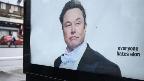 Getty Images A poster of Musk wearing a suit and bow tie on an advertising display panel in London, with "everyone hates elon" written on it. 