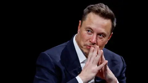 Reuters Elon Musk sitting down wearing a white shirt and navy blazer. He is looking to the left of the frame and has his hands clasped together in front of his mouth. He has short dark hair and stubble around his face.