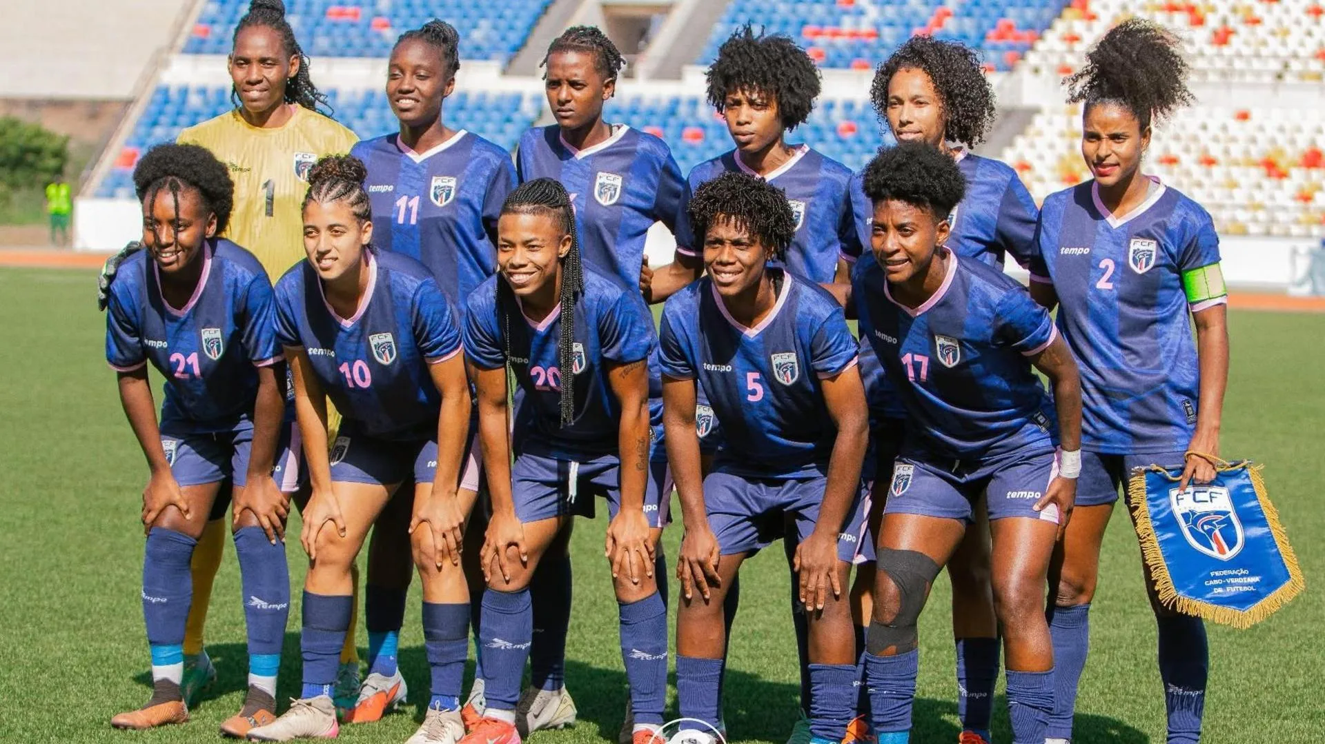 Cape Verde's women's footballers pose for a team photo before a game on a sunny day. Their goalkeeper, positioned far left of the back row, wears a golden jersey while the 10 outfield players are wearing an all-blue strip. The six players in the back row stand with arms around either other's waists, with the captain on the far right holding a national team pennant. The five players in the front row are standing but leaning forward with both hands on their knees