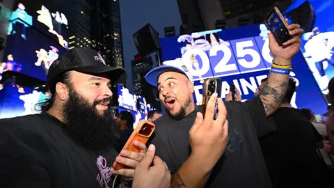 10 years of Pokémon Go and the millions still trying to catch 'em all AP Images for Scopely Two Pokémon Go players holding their phones up in the air as they play at night time in Times Square with an excited expression. There are bright large blue screens behind them and skyscrapers.