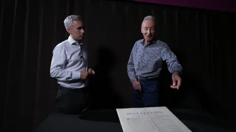 Tim Muffett and Michael Scurr stand in a dark room, with a copy of the US Declaration of Independence in front of them.