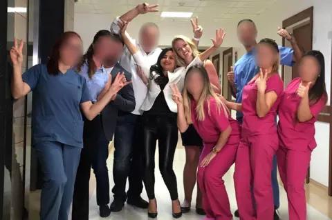 A group of 10 people stand together in a corridor, some wearing medical-style clothing. The group is posing with raised hands and gesture signs