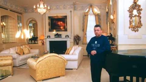 Shutterstock David Sullivan pictured at home in the 1990s, wearing a sweatshirt which says "The Blues" in reference to Birmingham City which he then co-owned. He is pictured leaning against a grand piano in an ornately decorated living room, with a crystal chandelier, gilt mouldings and mirrors, and sofas in white and gold.