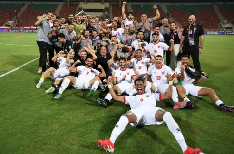 Ameen Ahmed/NurPhoto via Getty Images A group picture of Jordanian football players and other supporting members of the wider team smiling and celebrating on a football pitch.  Many of the players are wearing white kit and sitting on the ground. They are all looking at the camera.