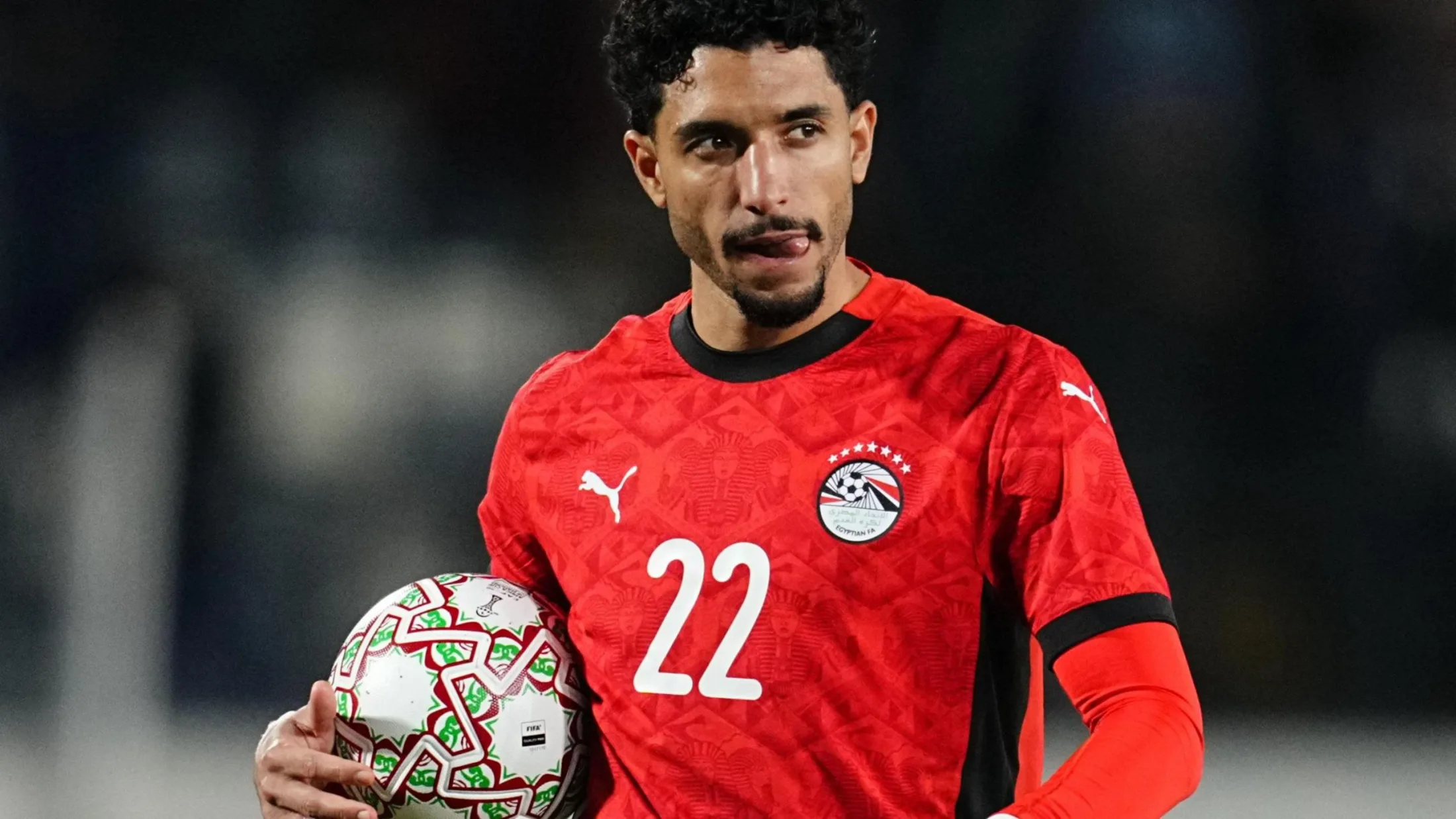 Omar Marmoush, visible from waist up wearing a red Egypt shirt with a white number 22 on the chest, sticks his tongue to the left side of his mouth as he cradles a football in between his right hand and his body
