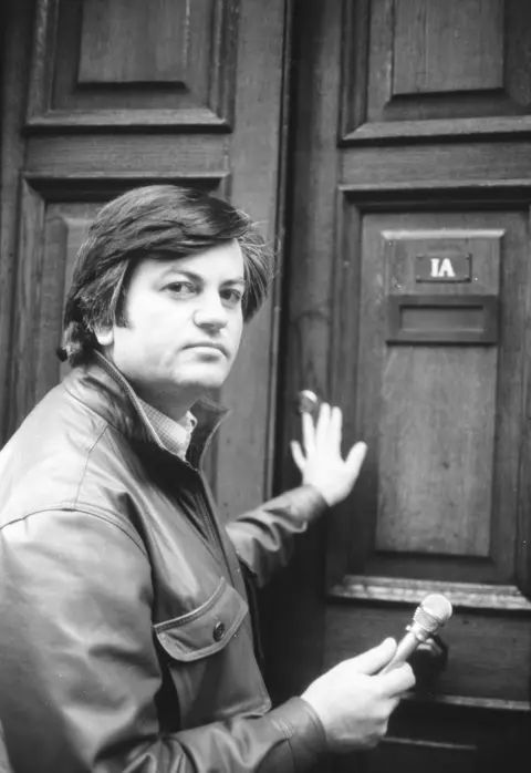 Roger Cook, pushing open a door, looking at the camera while holding a microphone