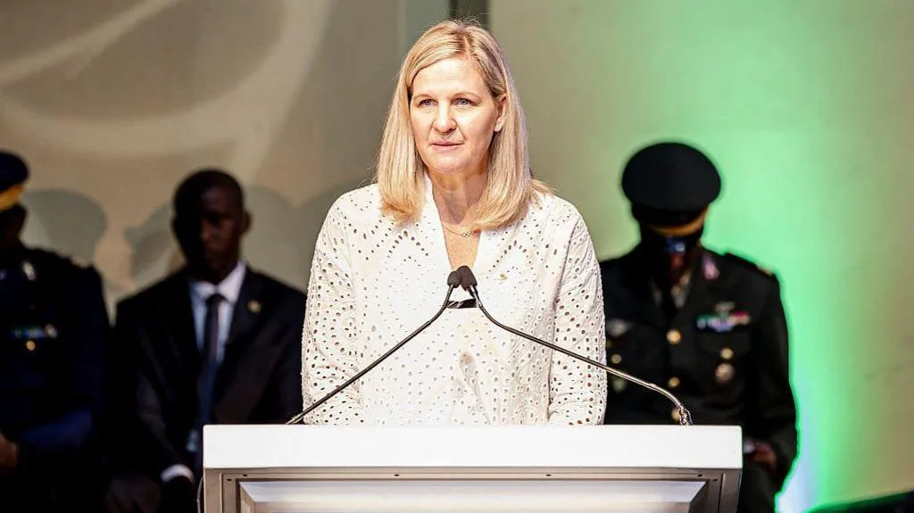 Kirsty Coventry, president of the International Olympic Committee, is seen wearing a white, long-sleeved blouse as she delivers a speech from a white lectern with two thin microphones positioned to meet in front of her. The background is softly blurred, showing several men in dark suits and military-style uniforms behind her under warm beige and green lighting.
