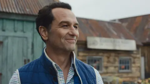 Apple TV Matthew Rhys in a striped shirt and blue body warmer in a still from Widow's Bay