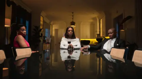The Polygamist's creator says women see themselves reflected in her Netflix hit Netflix/ Iconic Digital A tense dinner scene from the Netflix series reflected on a glossy black table: a woman in a pink dress sits at left, a woman in a white off-shoulder dress holds a wine glass in the center, and a man in a black tuxedo sits at right, all with serious, downcast expressions in a dimly lit, upscale kitchen/dining space.