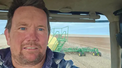 Geoff Cosgrove A man in a farming truck with a harvester in the background 