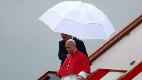 Pope says he will 'speak out loud' against war after Trump attack Reuters Pope Leo wearing papal cap and robes walks off a plane as a man in a dark suit behind him holds a white parasol over him.