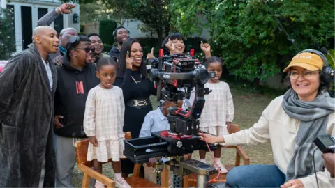 Netflix Behind the scenes at filming of The Polygamist showing some crew members and actors, including young twin girls standing on chairs in matching cheque dresses, smiling for a photo in a garden.