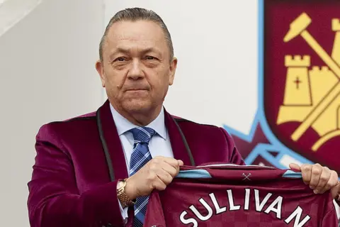 Getty Images David Sullivan, wearing a velvet jacket in West Ham's claret, holding a club shirt with his name written on the back, while standing in front of the club crest.
