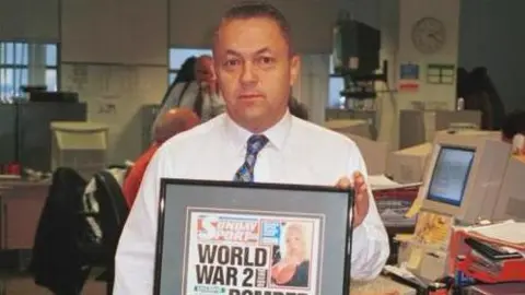 David Sullivan, pictured in 1999 for an episode of the BBC Two show Back to the Floor, in shirtsleeves with a tie, holding up a framed Sunday Sport front page with a blonde model in the top right corner and the headline: "World War 2 bomber found on moon". 