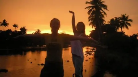 Lowri Denman Lowri and a friend dancing in the sunset in India. They stand in front of a lake with palm trees on the banks.