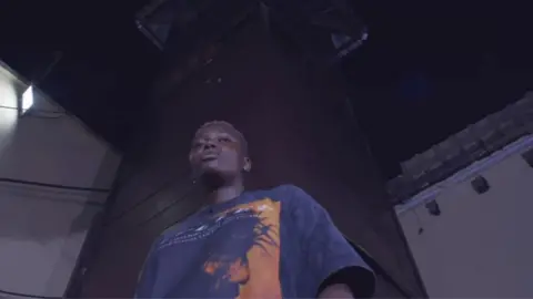 The notorious Cameroonian prison with its own record label Jail Time Records Jeje, a female prisoner, in a T-shirt standing in a prison yard with a guard tower behind her.