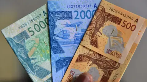 AFP via Getty Images Three CFA notes pictured - a green 5,000, a blue 2,000 and an orange 500 note.